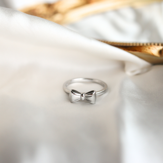 Silver bow-shaped ring on a white satin surface with a gold box in the background