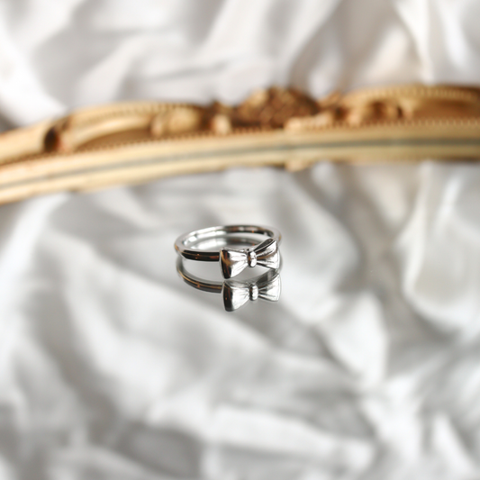 Silver bow-shaped ring on a textured white surface with a gold object in the background